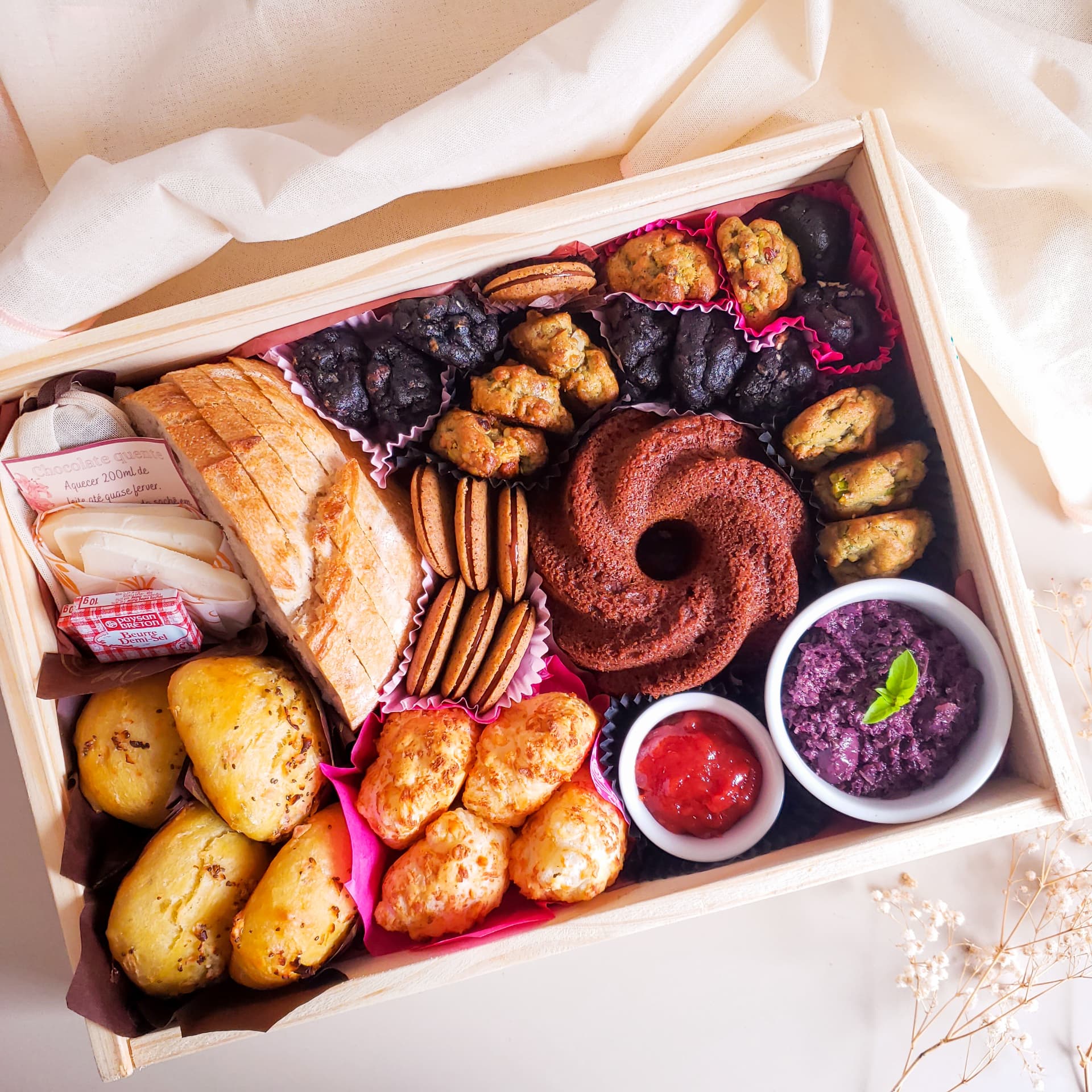 Caixa Gio da manhã aberta com fatias de pão, pãezinhos, pães de queijo, bolo, docinhos, dois ramequins e o preparo de bebida quente, com tecido de algodão cru e flores secas ao redor